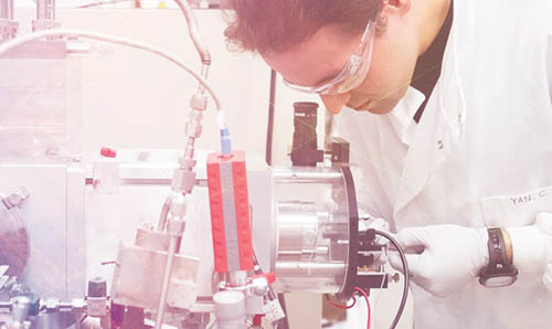 A male scientist in a white lab coat looks down a microscope.