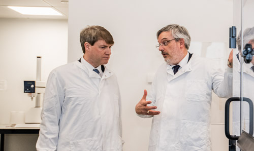 Two male scientists in white lab coats stand talking to each other.