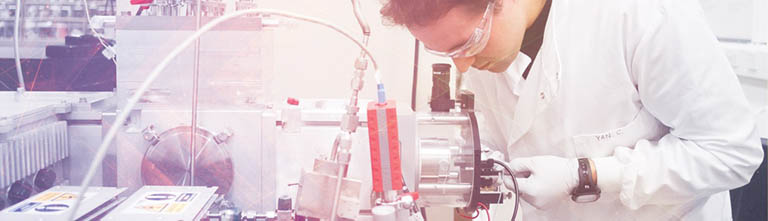 A male scientist wearing a white lab coat looks through a microscope.