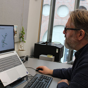 A scientist sits in front of a laptop that shows a chemical structure on the screen.