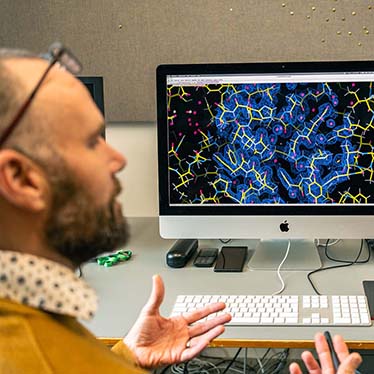 A man sits in front of a computer screen that is displaying a 3D render of a molecular structure.