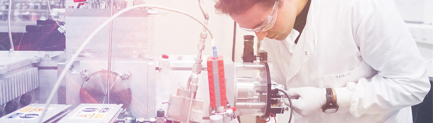 A male scientist in a white lab coat looks through a microscope